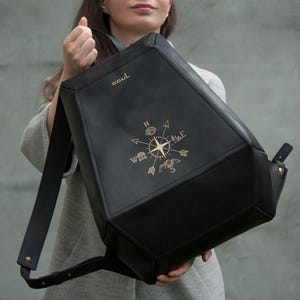May include: A black geometric backpack featuring a gold compass rose graphic on the front panel and the word SOUL printed in gold near the top. The bag has a structured, angular shape with wide black shoulder straps and gold metal hardware accents.