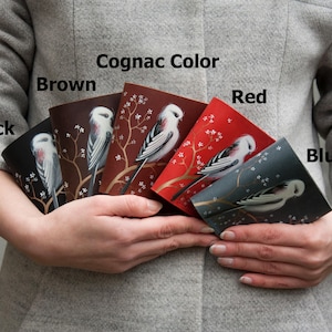 May include: Five rectangular leather covers in black, brown, cognac, red, and blue, each featuring a white bird perched on a brown branch with white blossoms. The text labels Black, Brown, Cognac Color, Red, and Blue are printed above each cover.