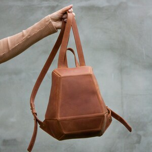 May include: A structured tan leather backpack with a geometric, multi-faceted design. The bag features a tapered body, a top handle, and adjustable shoulder straps with metal buckles. The surface has a smooth, matte finish with visible stitching.