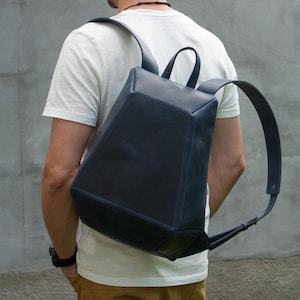 May include: A dark blue leather backpack with a structured, geometric, angular shape. The bag features a smooth surface, a top handle, and two adjustable shoulder straps. The design is minimalist with clean lines and a solid, deep navy color.