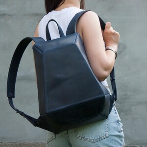 May include: A dark blue leather backpack with a structured, geometric, multi-faceted design. The bag features a top handle, two adjustable shoulder straps, and a wide, flat base. The surface has a smooth matte finish with visible stitching.