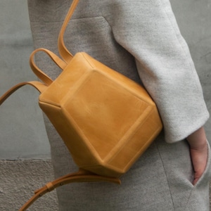 Puede incluir: Una mochila estructurada de cuero color amarillo mostaza con una silueta geométrica y cuadrada. El bolso presenta líneas limpias, costuras visibles a lo largo de los bordes y correas de hombro finas y ajustables fijadas en la parte superior e inferior.