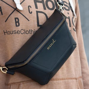 May include: Black leather fanny pack with a gold zipper and the word "SOUL" in gold lettering. The bag is worn across the body and has a gold clasp. The background is a brown hoodie with the text "HouseClothing".