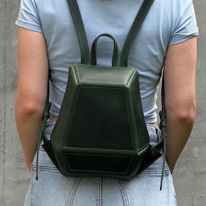 May include: A dark forest green backpack with a structured, geometric, multi-faceted shape. The bag features a top handle, two adjustable shoulder straps, and visible stitched seams along the edges of its angular, pentagonal-inspired panels.