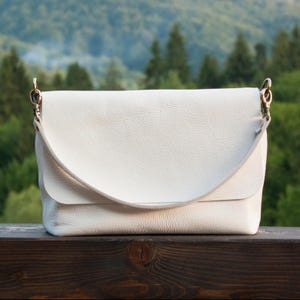 May include: A white leather crossbody bag with a flap closure and a shoulder strap. The bag has a textured surface and metal hardware. The background features a blurred view of green trees and a wooden surface.