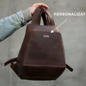 May include: A brown leather backpack with a unique geometric shape. The bag has a top handle and shoulder straps. The initials "EHN" are embossed on the front. The word "PERSONALIZATION" is written above the bag.