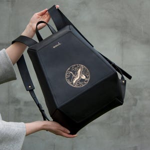 May include: A black geometric backpack featuring a gold-coloured circular emblem of a crane in flight. The word soul is printed in gold near the top edge. The bag has adjustable shoulder straps and a top handle, with a structured, angular design.