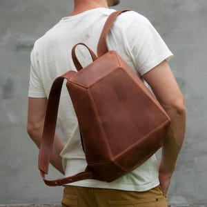 May include: A brown leather backpack with a geometric design. The backpack has adjustable straps and a top handle. The bag is worn over the shoulder. The person in the image is wearing a white t-shirt and brown pants.