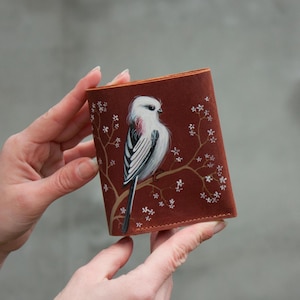 May include: A brown leather wallet featuring a hand-painted image of a long-tailed bird perched on a branch with white flowers. The bird has white, black, and pink feathers. The wallet is held by a person.