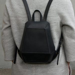 May include: A structured black backpack with a geometric, multi-faceted design. The bag features a top handle, two adjustable shoulder straps, and visible seam detailing along the edges of the main compartment, creating a distinct angular silhouette.