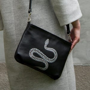 May include: Black crossbody bag featuring a white and grey snake design. The bag has a black strap and silver-coloured hardware. The snake illustration is detailed and prominent against the black background.