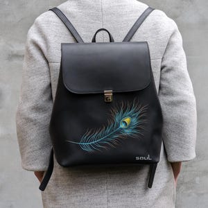 以下が含まれることがあります： 黒いバックパック。フラップ開閉とゴールドカラーの留め金付き。青、黄色、オレンジ色のカラフルな孔雀の羽のデザインが特徴です。「soul」の文字が羽の下部に白で印刷されています。