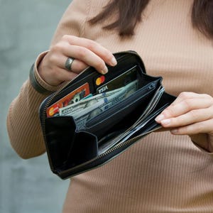 May include: Black leather wallet with a zipper closure, open to reveal cash, credit cards, and various compartments. The wallet is held open, showcasing its contents. The interior features multiple card slots and bill compartments.