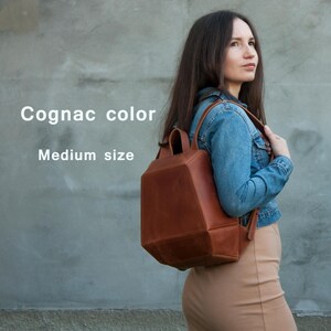 May include: A medium size cognac color leather backpack with a structured geometric shape, featuring a top handle and two shoulder straps. The text Cognac color and Medium size is printed on the left side of the image in white lettering.
