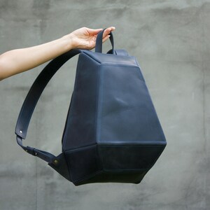 May include: A dark navy blue backpack with a geometric design. The backpack has a handle and adjustable straps. The bag is held up by a person's hand. The background is a solid gray color.