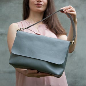 May include: A grey leather shoulder bag with a flap closure and a long strap. The bag features gold-coloured hardware and a textured surface. The bag is held up by the strap.