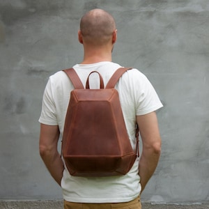 May include: A structured brown leather backpack with a geometric, multi-faceted design. The bag features a top handle, two wide shoulder straps, and a tapered silhouette that narrows toward the top, showcasing smooth leather panels and visible stitching.