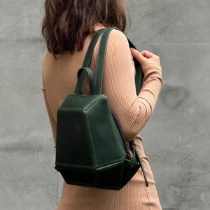 Pode incluir: Uma mochila estruturada em verde floresta escuro com um design geométrico de múltiplas facetas. A bolsa possui uma alça superior, duas alças de ombro ajustáveis e um fecho de correr lateral. O exterior tem um acabamento liso e mate.