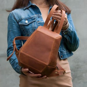 May include: A brown leather backpack with a geometric design. The backpack has two shoulder straps and a top handle. The bag is held by a person wearing a denim jacket.