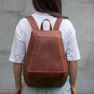 May include: A structured brown leather backpack with a geometric, multi-paneled design. The bag features a top handle, two adjustable shoulder straps, and visible stitching along the edges of the angular, tapered body construction.
