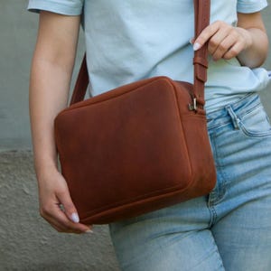 Könnte beinhalten: Eine rechteckige, rostfarbene Umhängetasche aus Leder mit einer glatten, matten Oberfläche. Die Tasche verfügt über einen Reißverschluss an der Oberseite, einen passenden verstellbaren Schulterriemen aus Leder mit Metallschnalle und Nähte.