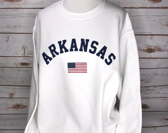Può includere: Felpa bianca con un arco blu navy che recita "ARKANSAS" e una piccola bandiera americana al centro.