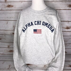 May include: A gray sweatshirt with the text "ALPHA CHI OMEGA" in blue lettering in an arc above a small American flag.