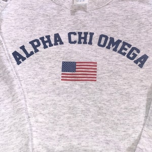 May include: Gray sweatshirt with the text "ALPHA CHI OMEGA" in blue letters in an arc above a small American flag.