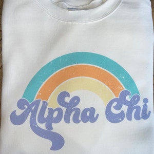 May include: White sweatshirt with a rainbow design and the text "Alpha Chi" in a retro font.