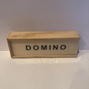 May include: A wooden box with the word "DOMINO" printed in black capital letters. The box is rectangular and appears to be made of light-colored wood. The box is closed and ready for play.