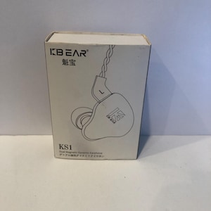 Puede incluir: Una caja blanca con la marca "KBEAR" y el modelo "KS1" impresos. La caja presenta un dibujo lineal de auriculares internos blancos con un cable plateado. El texto "Dual Magnetic Dynamic Earphone" también está impreso en la caja.