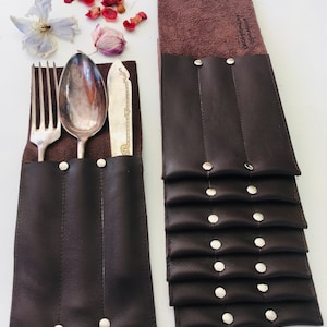 Può includere: Set di portaposate in pelle marrone con bottoni a pressione argentati. Ogni portaposate ha tre fessure per una forchetta, un coltello e un cucchiaio. I portaposate sono impilati uno sopra l'altro.