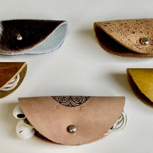 Può includere: Cinque piccole custodie per auricolari in pelle fatte a mano in vari colori e trame. Le custodie sono fissate con bottoni automatici in metallo e contengono auricolari e cavi. Una custodia è in pelle di mucca e un'altra ha un motivo decorativo.