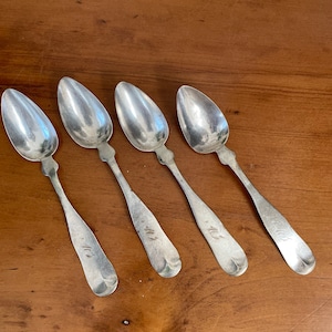 May include: Four silver spoons with ornate handles. Each spoon has an inscription on the back of the handle. The spoons are arranged in a row on a wooden surface.