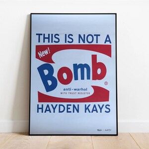 May include: A blue and red poster with the text "THIS IS NOT A BOMB" and "New!" in a white box. The word "Bomb" is in a large blue font with a red outline. The text "anti-warhol WITH TRUST RESISTER" is in a smaller white font. The poster is signed "Hayden Kays" in the bottom right corner.
