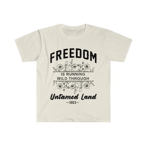 Könnte beinhalten: Cremefarbenes T-Shirt mit dem Wort "FREEDOM" über einem floralen Design und dem Text "IS RUNNING WILD THROUGH UNTAMED LAND -1883-" in Schwarz. Das Shirt hat kurze Ärmel und einen Rundhalsausschnitt.