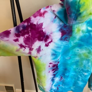 Può includere: Una felpa con cappuccio tie-dye con uno schema di colori blu, verde, viola e bianco. La felpa ha un cappuccio e un cordoncino.