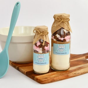 May include: Two glass jars filled with s'mores cookie mix ingredients. The jars are tied with twine and have brown paper labels that say "S'mores Cookie Mix".