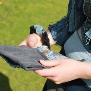 May include: A person is holding a folded piece of dark grey fabric in their hands. The fabric is folded in half and the edges are visible. The person is wearing a denim jacket and jeans.