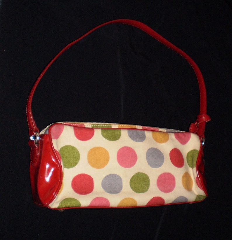 May include: A red and white polka dot handbag with a red leather strap. The bag has a zip closure and a small pocket on the front.