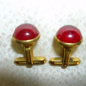 May include: A pair of gold cufflinks with red glass stones. The cufflinks have a simple design with a round stone set in a gold frame.