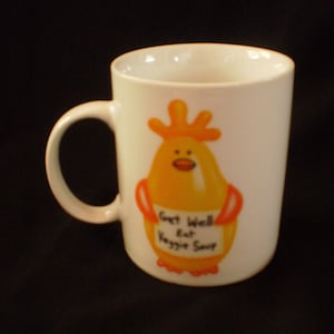 May include: White ceramic mug featuring a cartoon chicken with orange and yellow coloring. The chicken holds a sign that reads "Get Well Eat Veggie Soup". The mug has a curved handle.