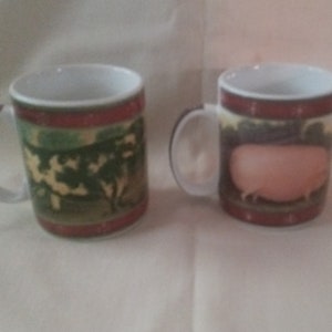 May include: Two ceramic mugs with white handles and interiors. One mug features a landscape scene with trees, the other a pink pig. Both mugs have a red band at the top and bottom.