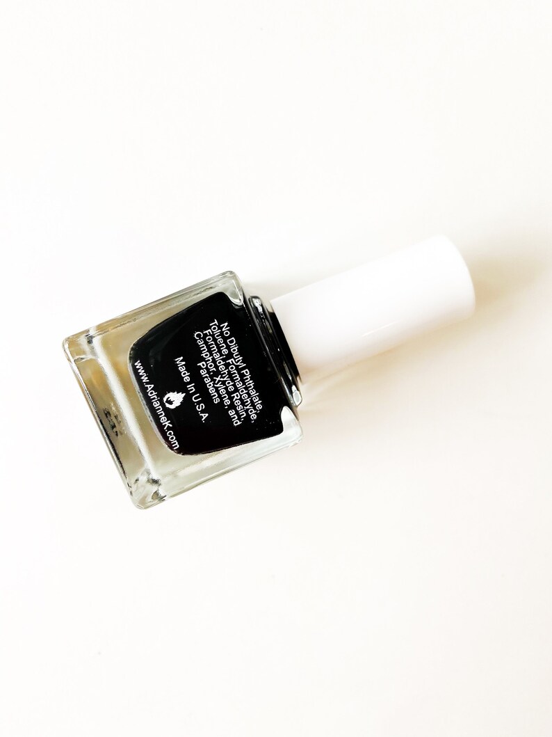 May include: A clear glass bottle of black nail polish with a white cap. The bottle label lists ingredients and states "No Dibutyl Phthalate, Toluene, Formaldehyde, Formaldehyde Resin, Camphor, Xylene, and Parabens. Made in U.S.A. www.Adriannek.com"