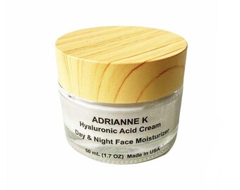 May include: A white jar with a wooden lid containing a hyaluronic acid cream. The jar is labeled "ADRIANNE K Hyaluronic Acid Cream Day & Night Face Moisturizer 50 mL (1.7 OZ) Made in USA."