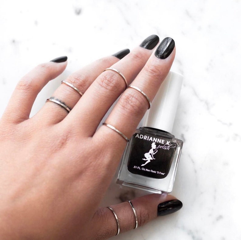 May include: A hand with black nail polish on the index finger, wearing several silver rings, holding a bottle of black nail polish labeled "Adrianne K polish".
