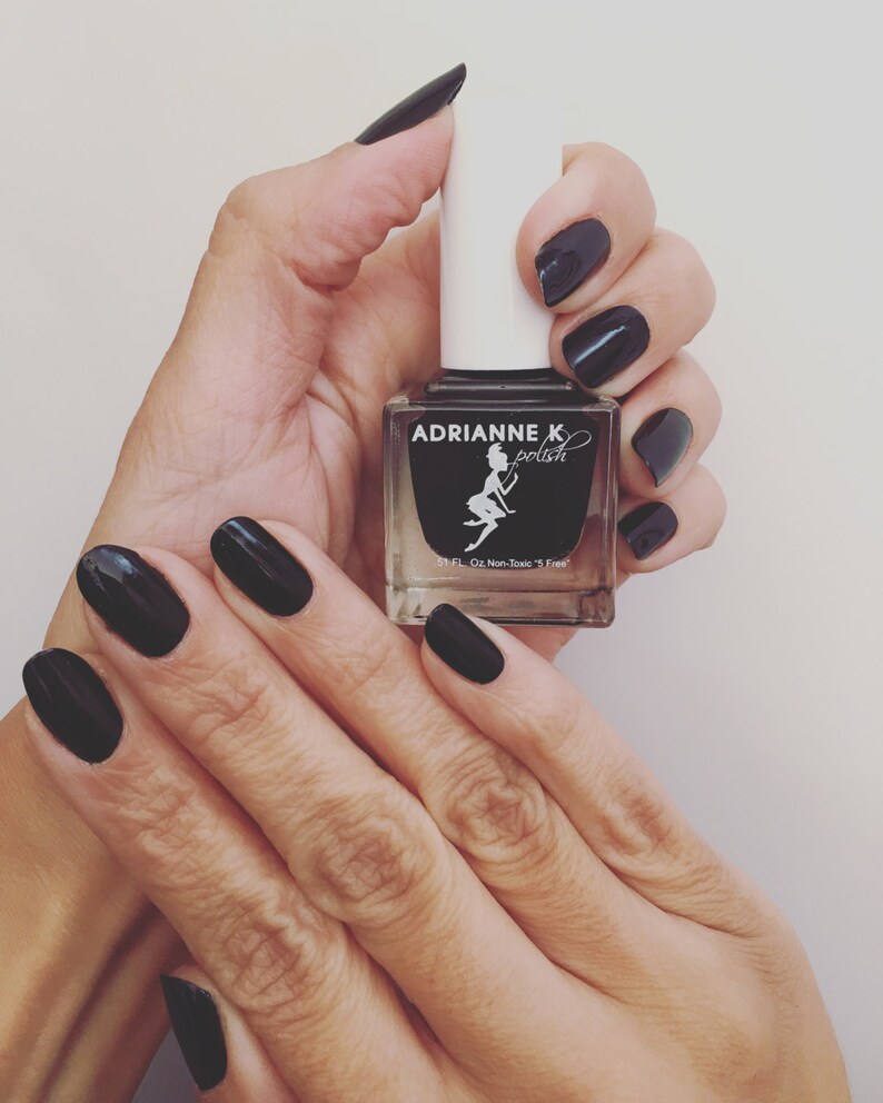 May include: A black nail polish bottle with the brand name "Adrianne K polish" and a fairy illustration. The bottle is being held by a hand with black nail polish.