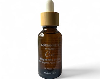 Op de afbeelding: Een amberkleurige glazen druppelfles met een licht houten dop en een witte rubberen knijptuit. Het etiket vermeldt ADRIANNE K VITAMIN C+E Brightening Serum With Organic Apple Extracts, 30 ml, Made in USA. De fles bevat een heldere vloeistof.