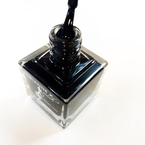 May include: A square bottle of black nail polish with a silver cap and a black brush. The bottle is labeled "Adrianne Special Non-toxic".