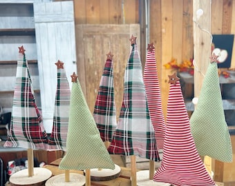 May include: A collection of decorative fabric Christmas trees in various patterns, including plaid, polka dots, and stripes. Each tree is mounted on a wooden base and topped with a wooden star. The trees are displayed on a wooden surface.